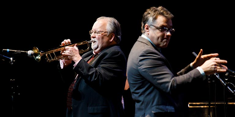 A conductor conducts an orchestra as a man plays the trumpet.