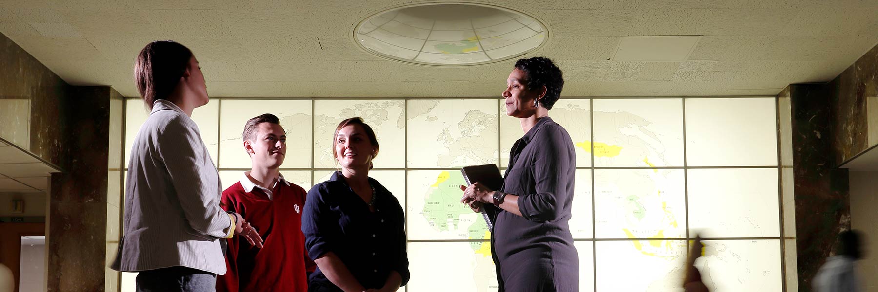Three students talk with a professor in front of a lighted wall showing a world map.